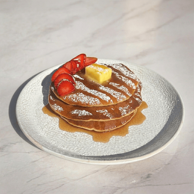 Pancakes Clásicos Artesanales - Tres esponjosos pancakes dorados, coronados con mantequilla fundente, fresas frescas laminadas y una delicada lluvia de azúcar flor, bañados con auténtica miel de maple. Un equilibrio perfecto entre suavidad, frescura y dulzor. en Le Grenier